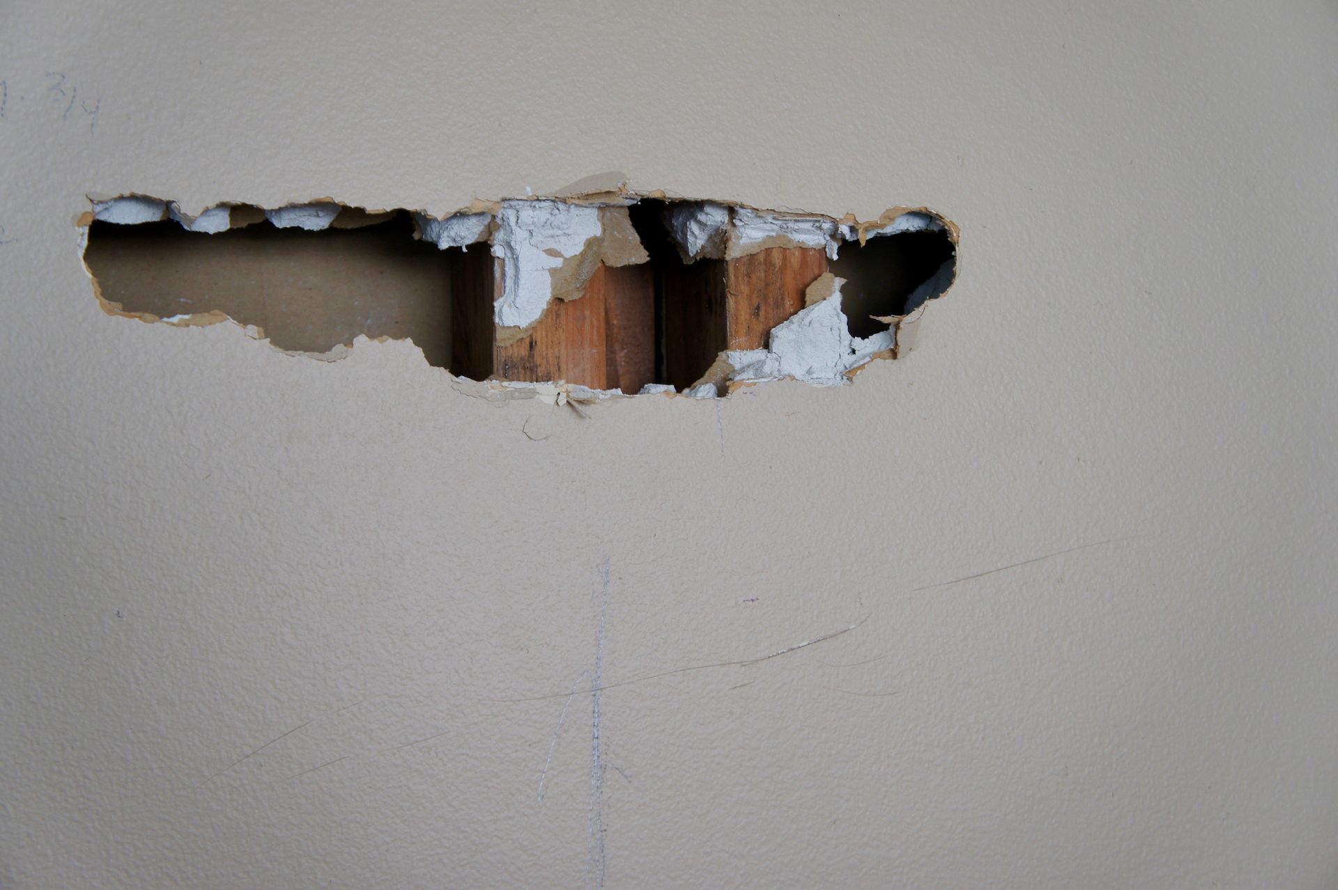 Hole in a tan wall revealing wooden studs. White paint chips surround the opening.