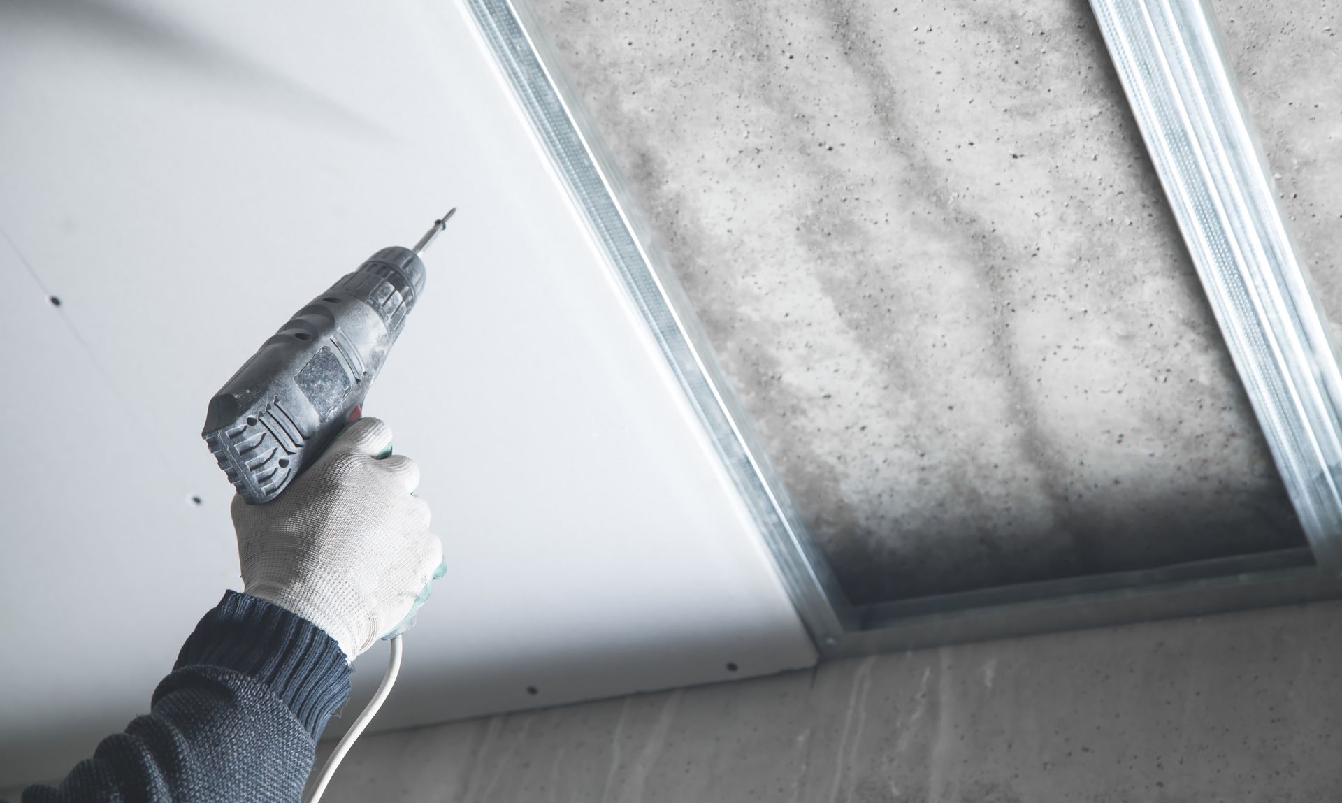 Person in gloves using a drill to install drywall on a ceiling.