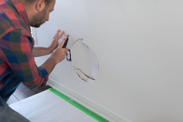 Man crouching down and repairing a dent on a white drywall near a electric outlet.