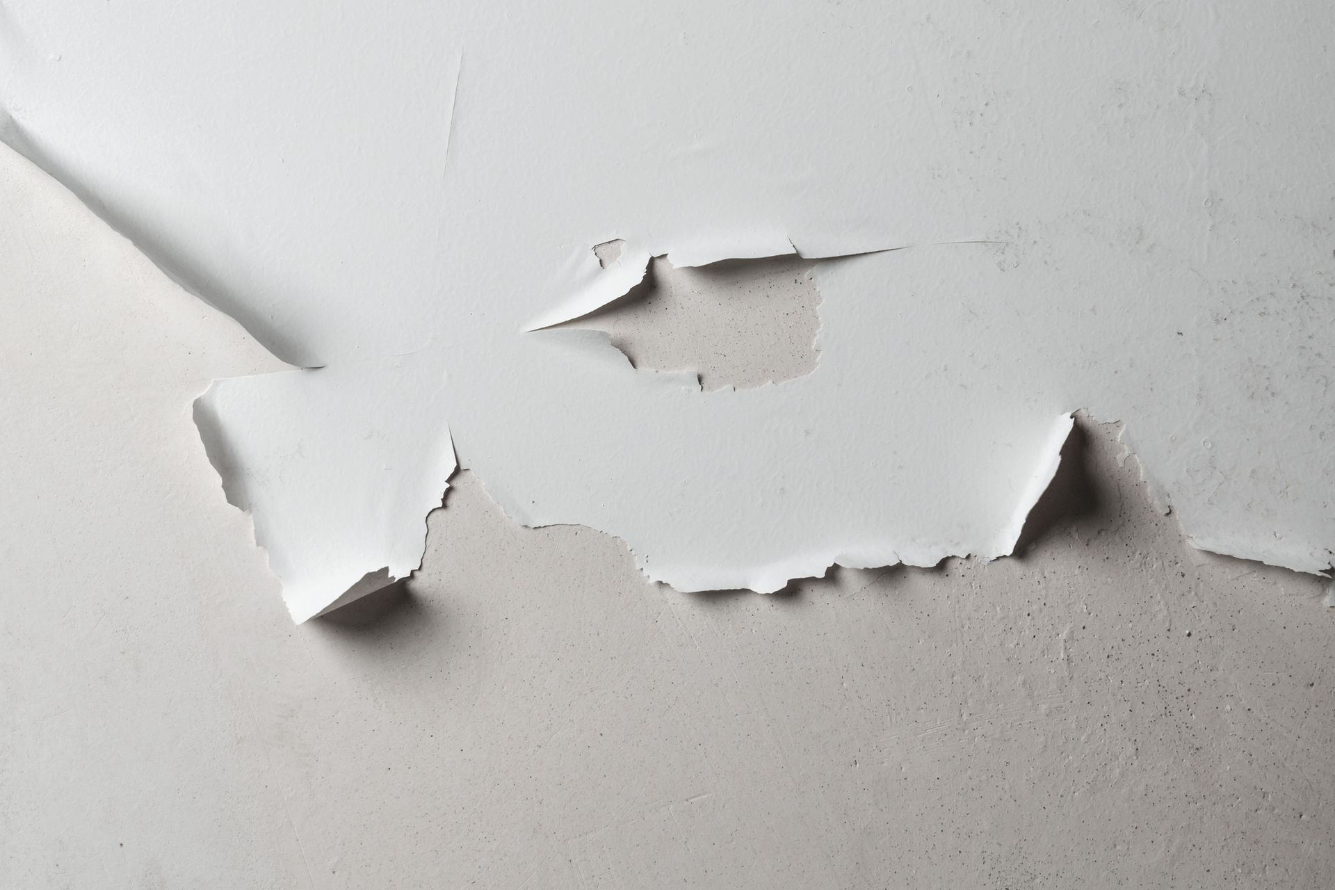 Flaking white paint on a light gray wall, revealing the surface beneath. Flaking white paint on a light gray wall, revealing the surface beneath.
