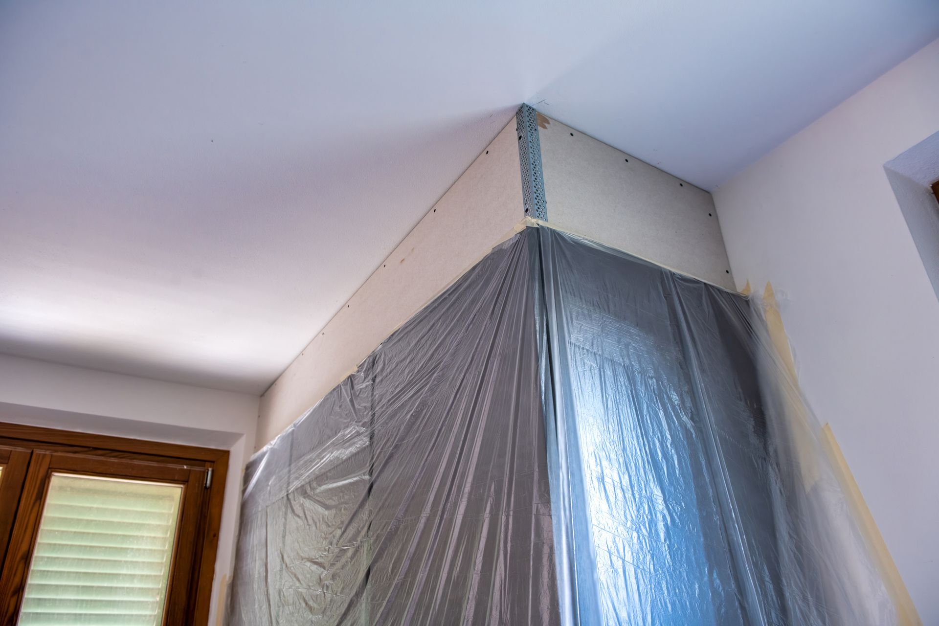 Corner of a room under renovation. Drywall, plastic sheeting, and a window frame are visible. Corner of a room under renovation. Drywall, plastic sheeting, and a window frame are visible.