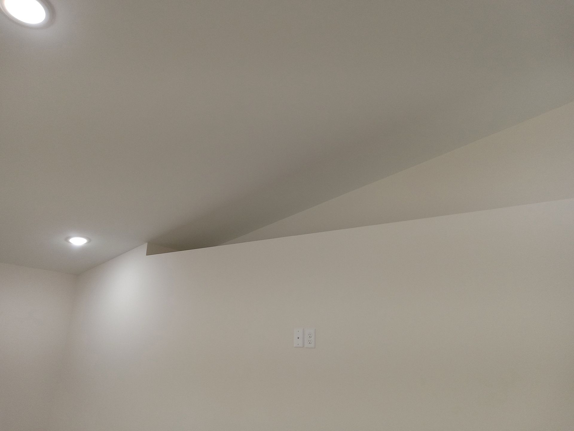 White room with angled ceiling and wall meeting. Recessed lights are visible. White room with angled ceiling and wall meeting. Recessed lights are visible.