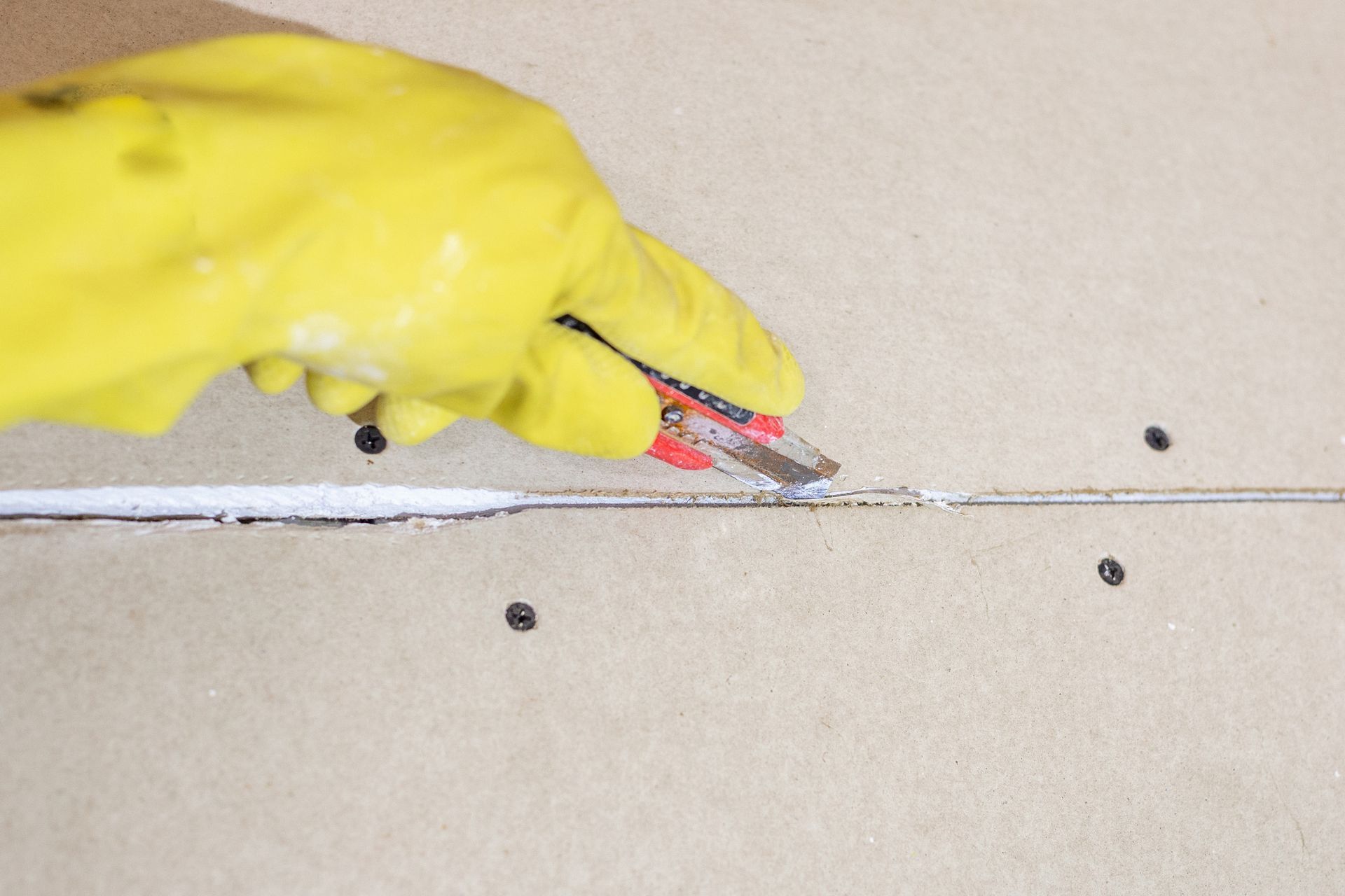 Gloved hand using a utility knife to cut dried caulk between two wall panels. Gloved hand using a utility knife to cut dried caulk between two wall panels.