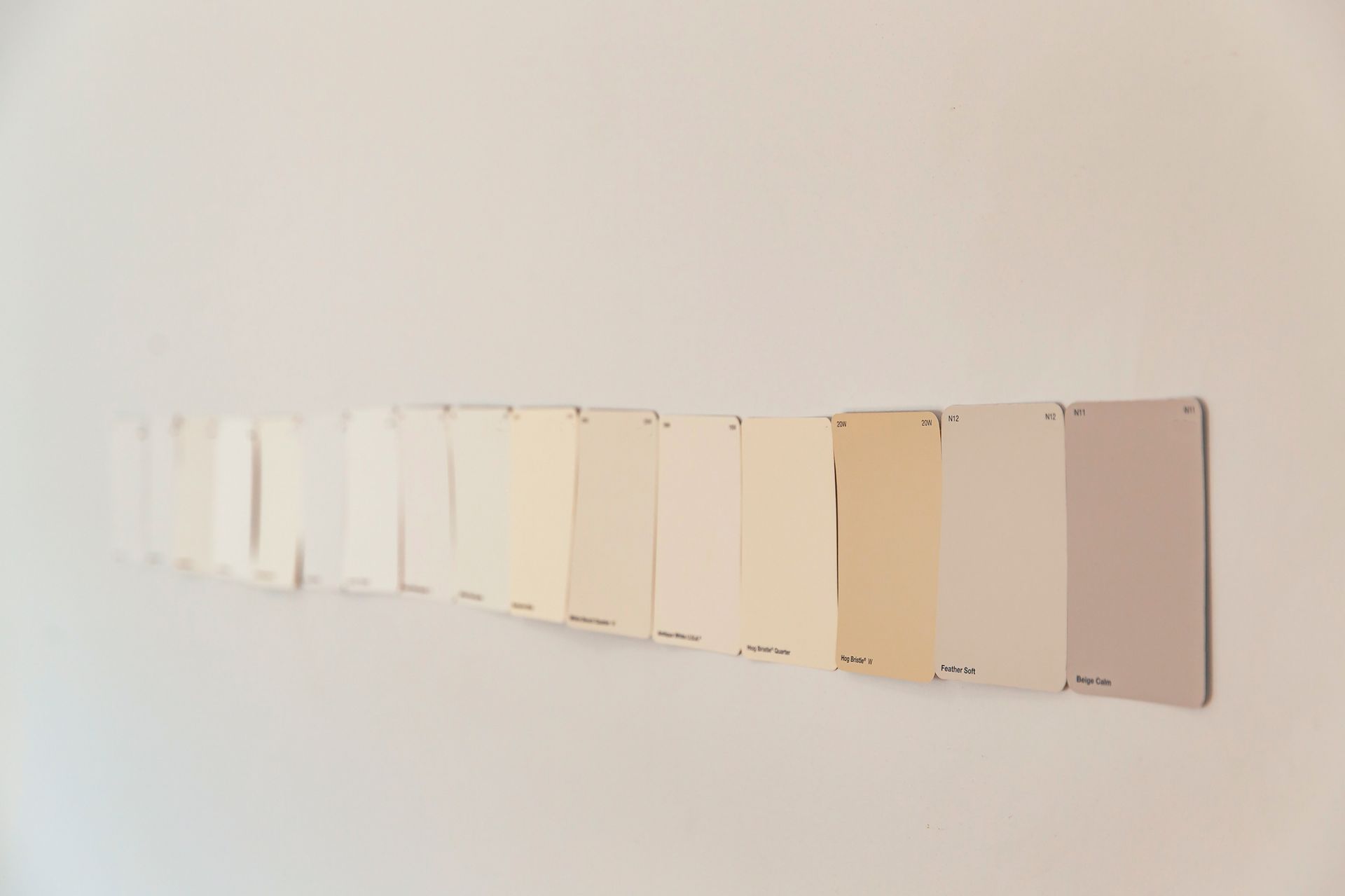Paint color swatches in a gradient from white to beige and light purple, attached to a white wall. Paint color swatches in a gradient from white to beige and light purple, attached to a white wall.