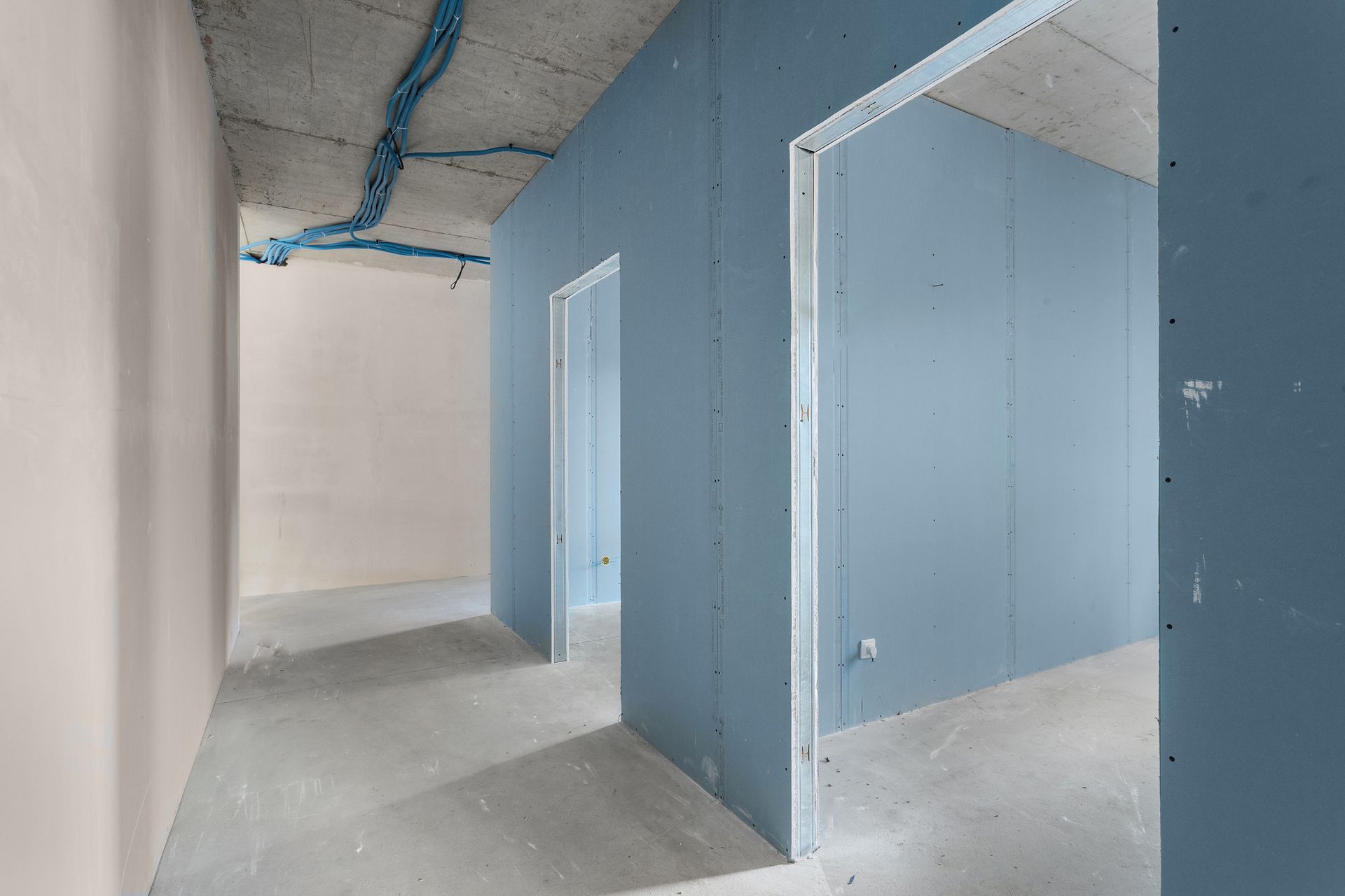 Interior hallway in construction phase, with unfinished walls and doorways, blue drywall, and concrete floors. Interior hallway in construction phase, with unfinished walls and doorways, blue drywall, and concrete floors.
