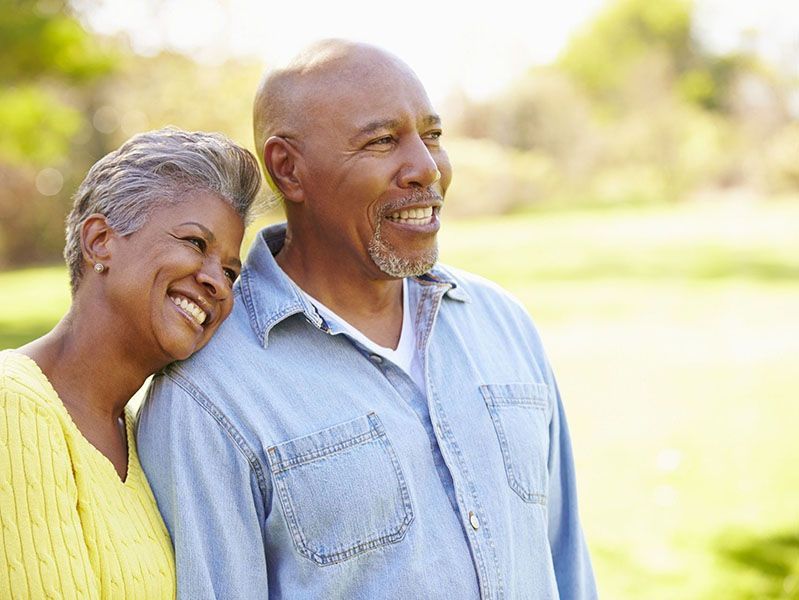 Smiling couple outdoors, woman leaning on man. Both looking off to the side, sunny background.