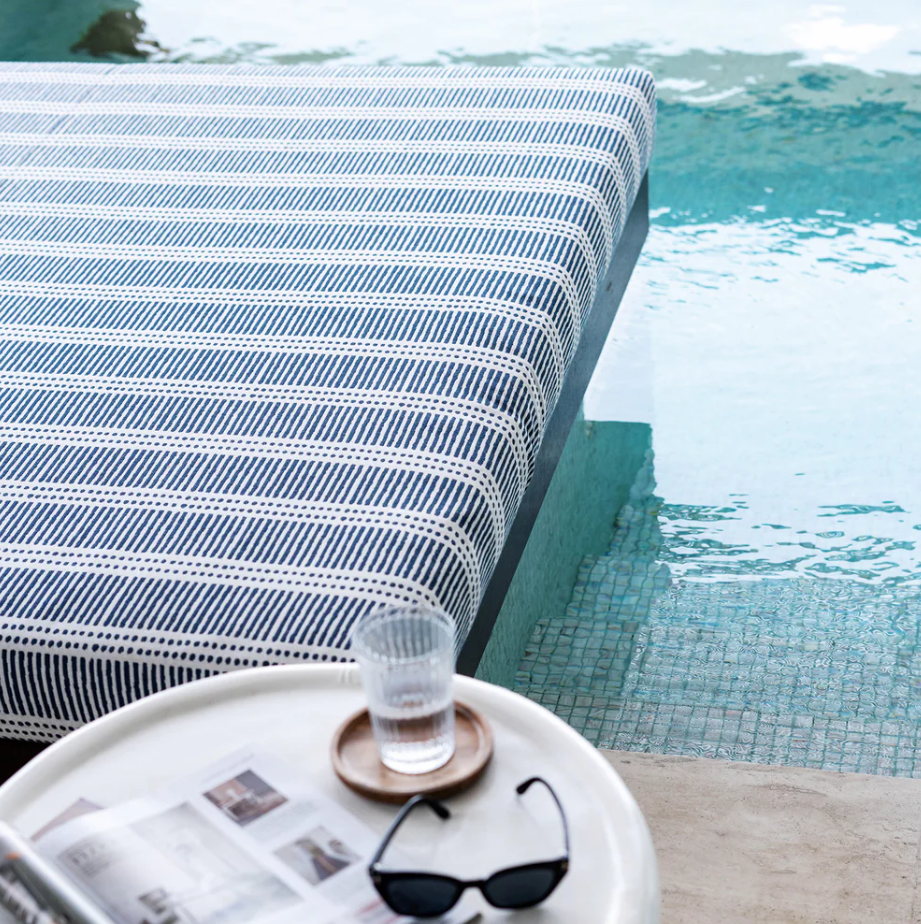A blue and white striped daybed sits beside a swimming pool, with sunglasses and a drink on a nearby table.