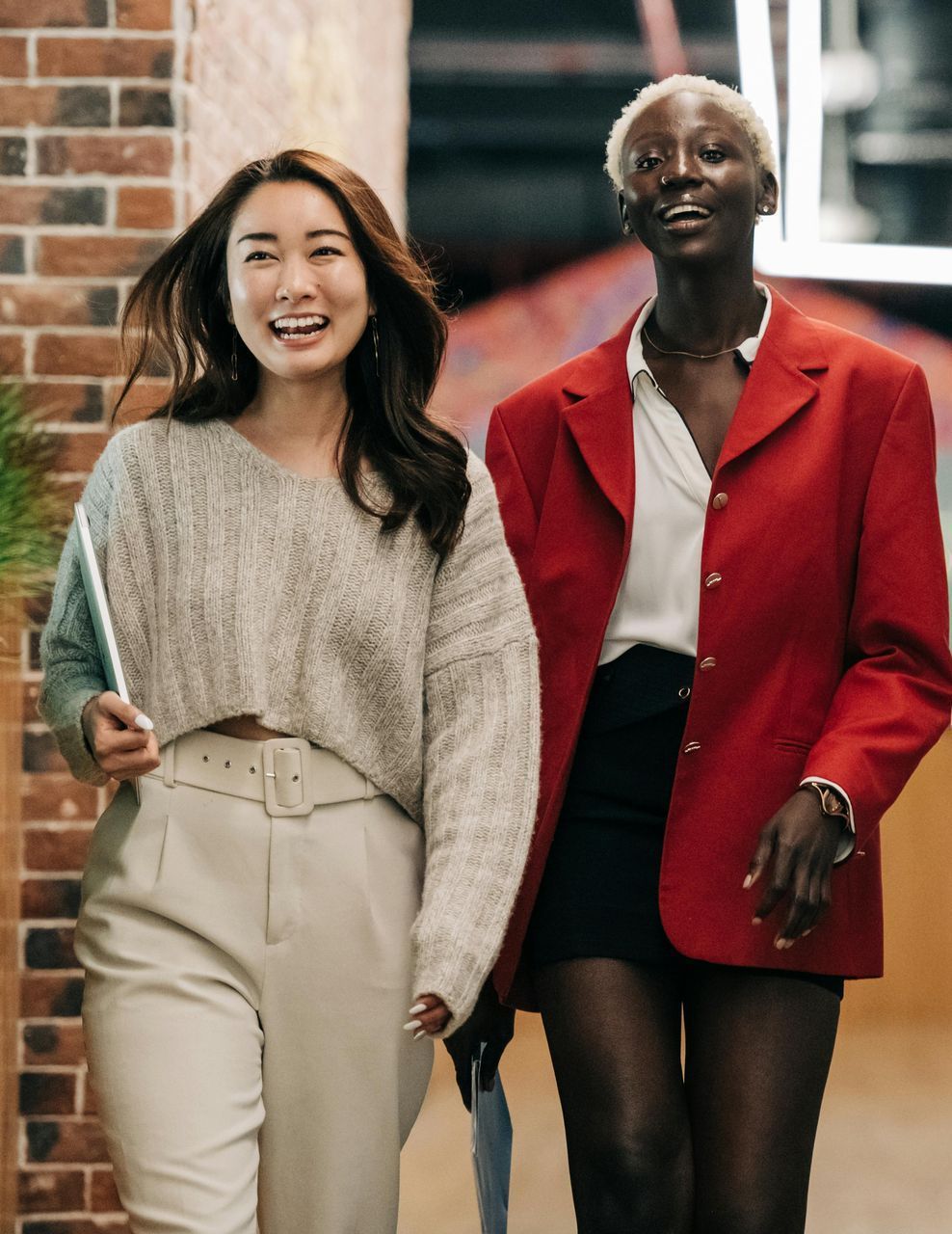 Two women are walking down a hallway and one is wearing a red jacket.