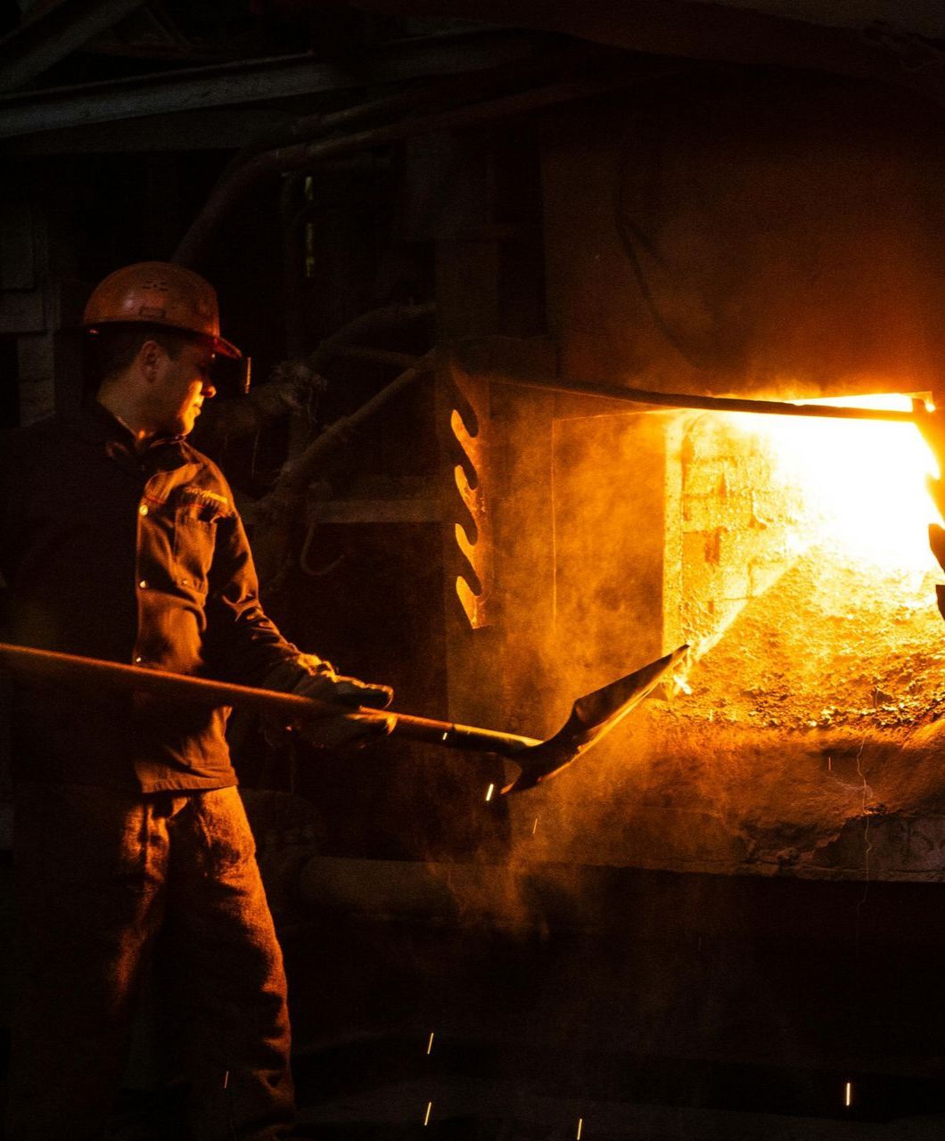 A man wearing a hard hat is holding a shovel in front of a fire