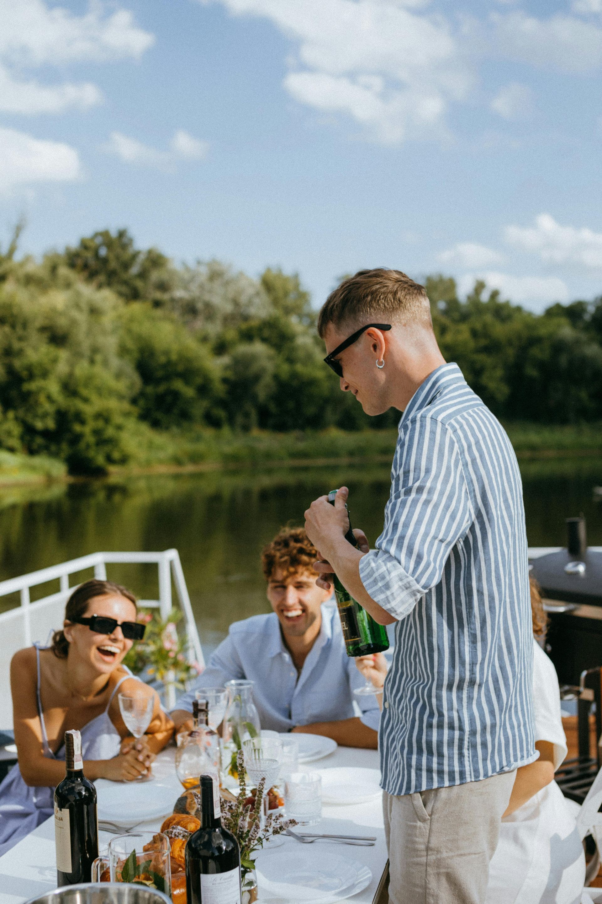 A man is pouring wine into a bottle while a group of people are sitting at a table.