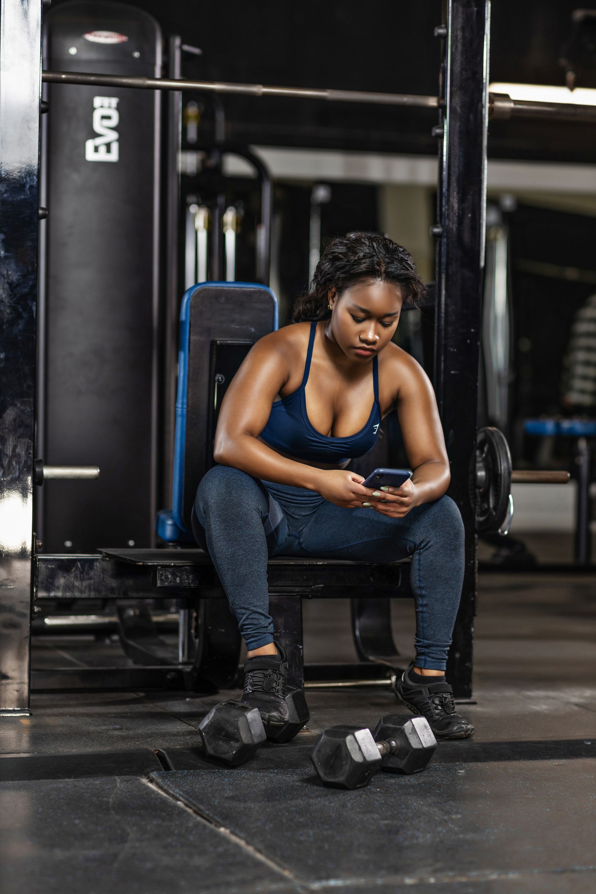 Woman in workout clothes, using a phone while resting on gym equipment with dumbbells on the floor. B for Body