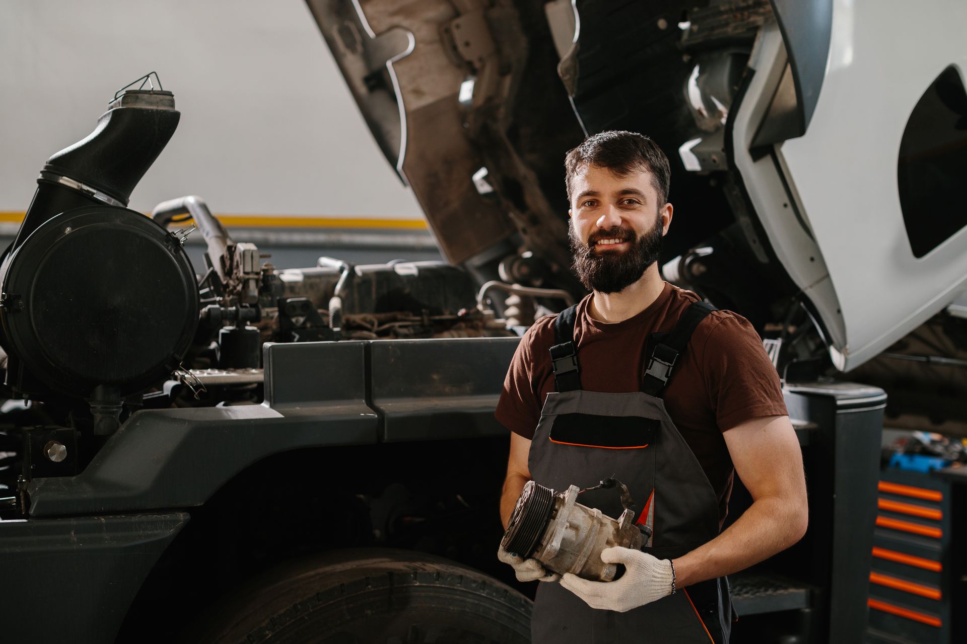 Mechanic holding engine part near a truck with open hood in a workshop.