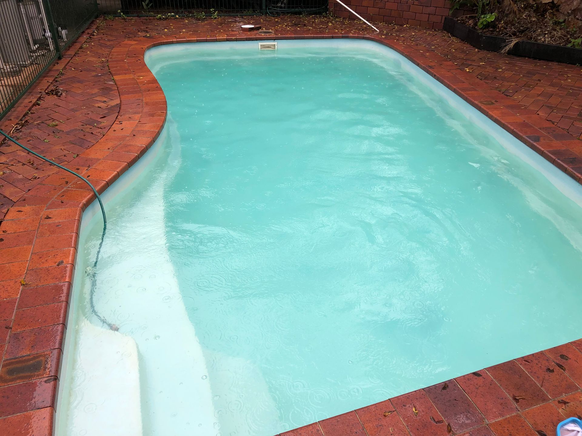 Light blue swimming pool surrounded by wet red brick paving, with a hose filling the water.