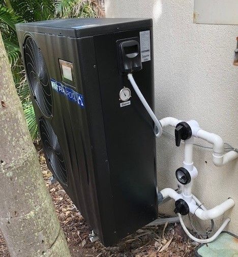Black Pool Heater Next To A Wall With White Pipes And Valves. Outdoors With A Tree In The Background