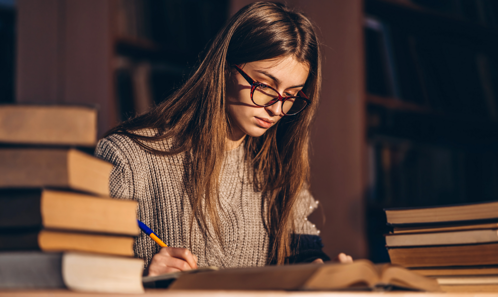 A woman wearing glasses is sitting at a table with a stack of books.