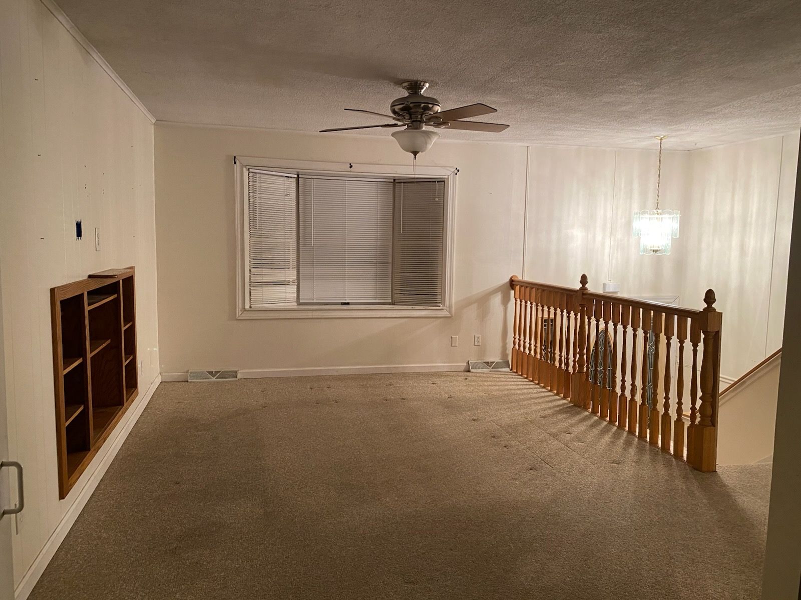 A carpeted room with a wooden railing, built-in shelving, a window with blinds, and a ceiling fan.