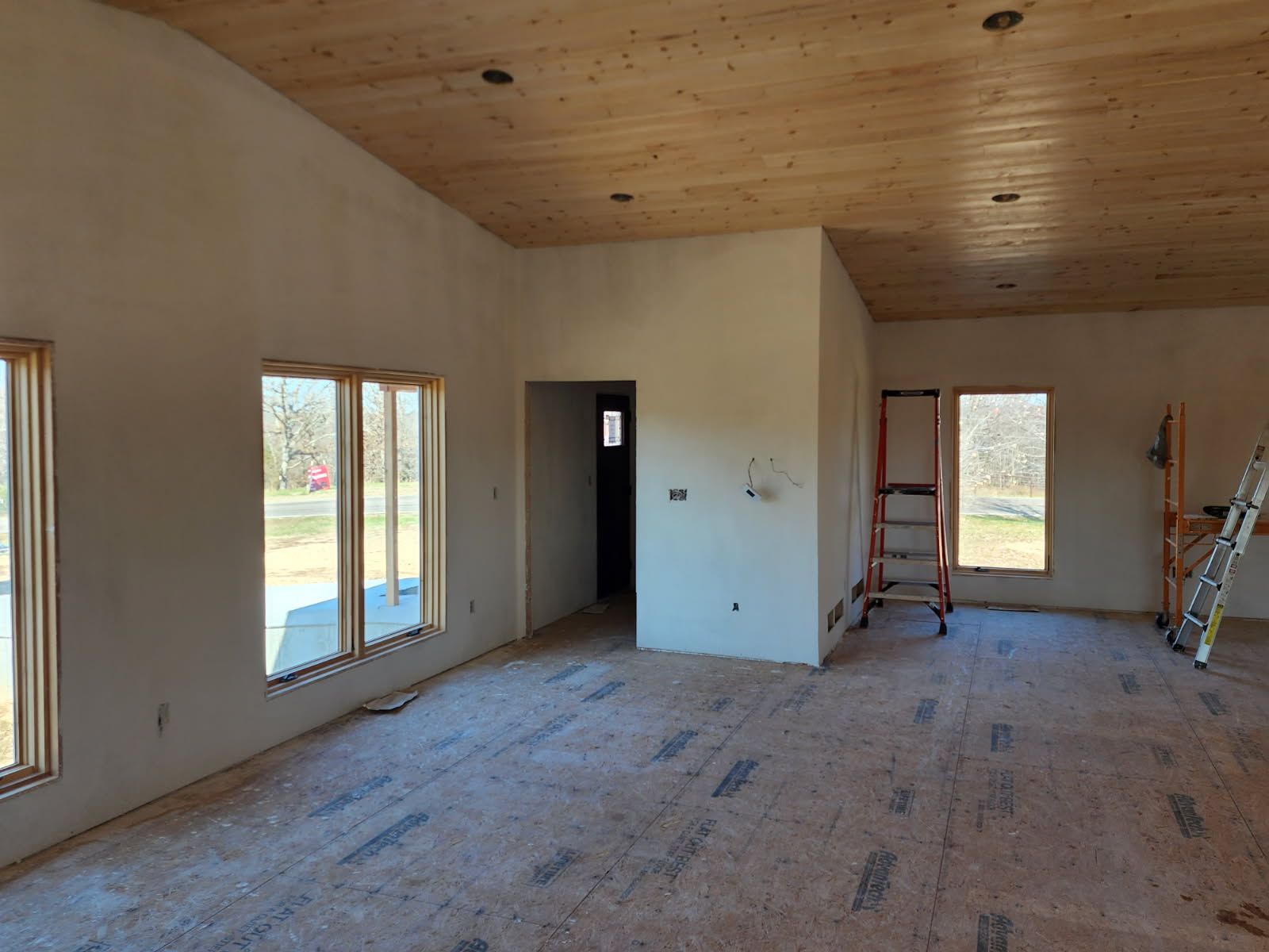 Interior of a room under construction, with wooden ceiling, bare walls, windows, door frame, and ladders.