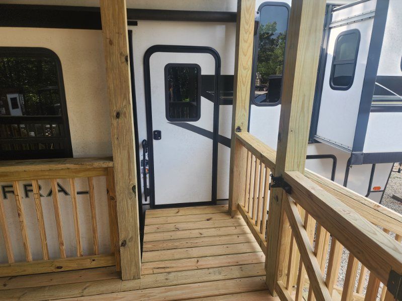 Wooden porch with door leading to a white RV, with railings, and partially visible RV windows.