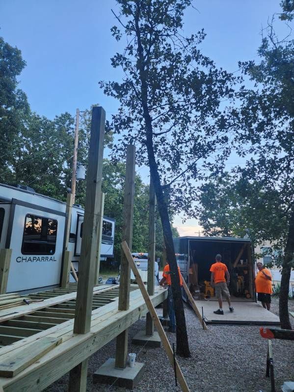 Construction of a deck next to a white RV, with workers in orange shirts. Tree is between the deck and RV.