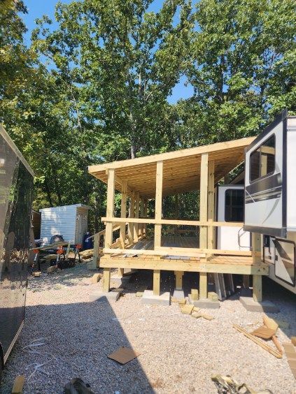 A wooden porch being built next to a camper; construction in progress outdoors, trees in the background.