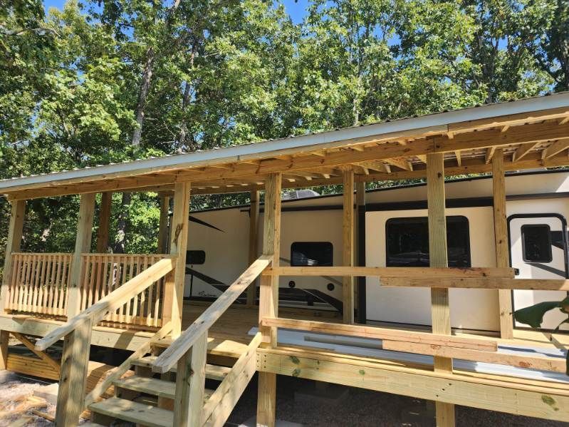 Wooden deck and porch attached to a white RV, set in a wooded area.
