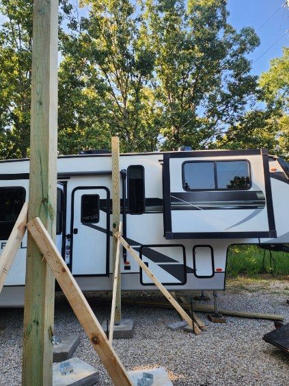Wooden support posts braced near a white RV parked outdoors, gray gravel, trees in background.