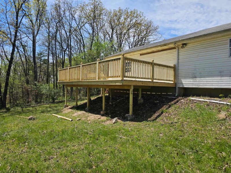 Wooden deck built on posts, attached to a light-colored house. Green grass and trees surround it.
