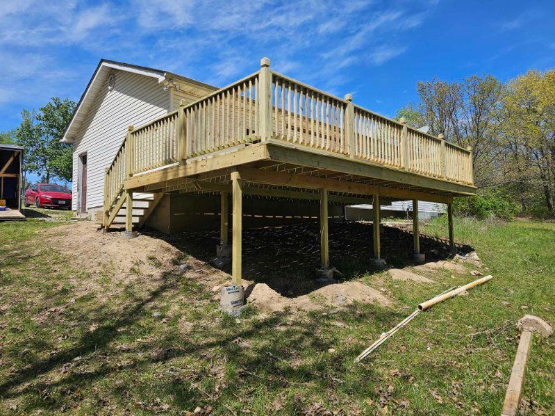 Newly built wooden deck attached to a house with stairs, set on posts, and a railing on a sunny day.