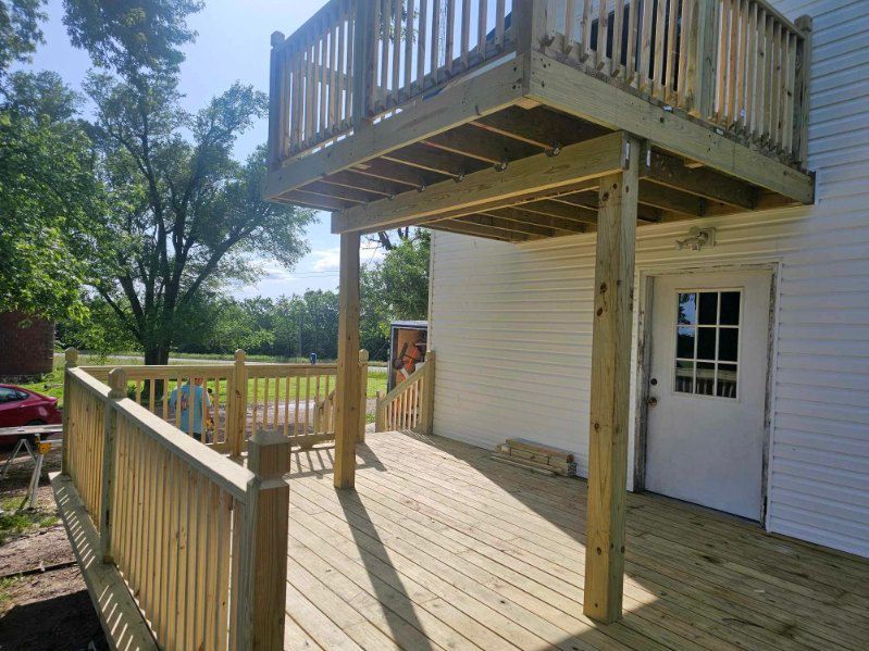 Wooden deck and balcony attached to a white house, supported by wooden posts. Sunny outdoor scene.