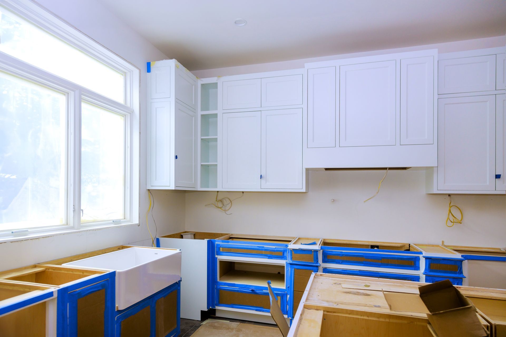 Kitchen under construction with white cabinets, blue tape, and window.