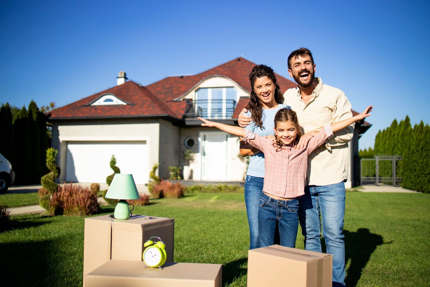 Family of three stands in front of a new house, excited, with moving boxes on the lawn.