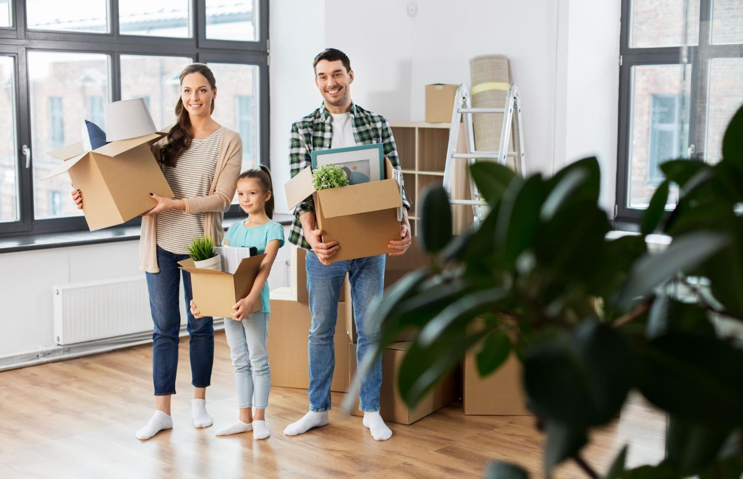 A family standing in a sunny, unfurnished room, smiling while holding cardboard moving boxes.
