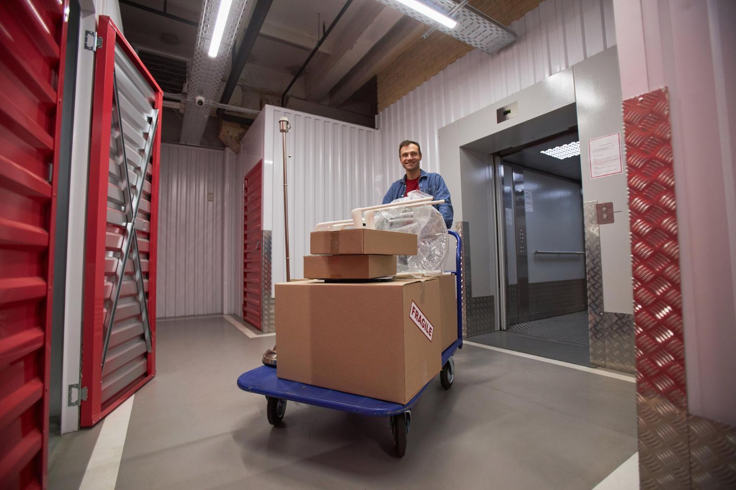 Man with boxes on a cart in a storage facility corridor, near elevator and open storage units.