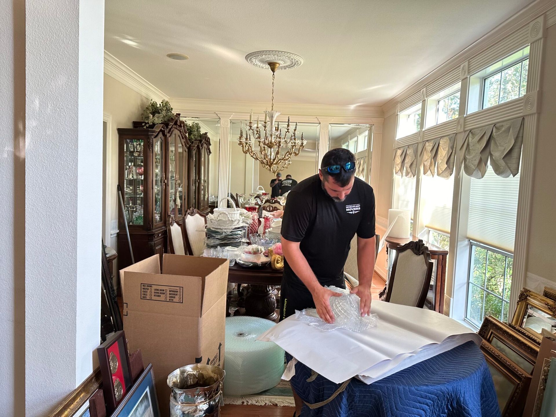 A man wraps items in a dining room with a chandelier and packed boxes.