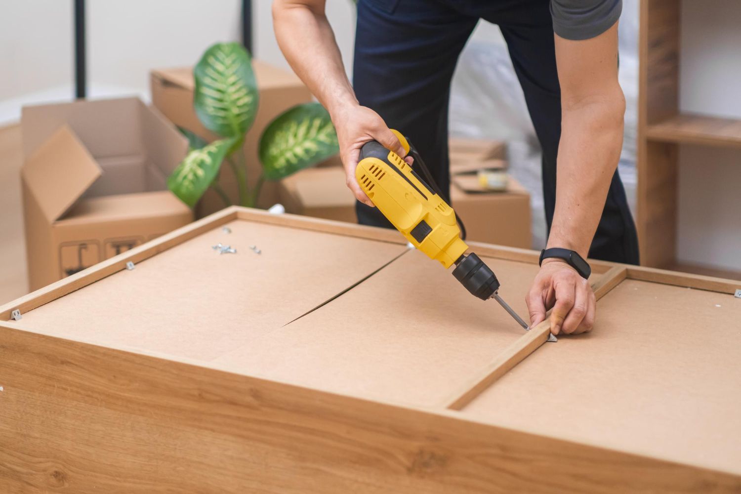 Person assembling furniture with a yellow power drill inside a room with moving boxes.
