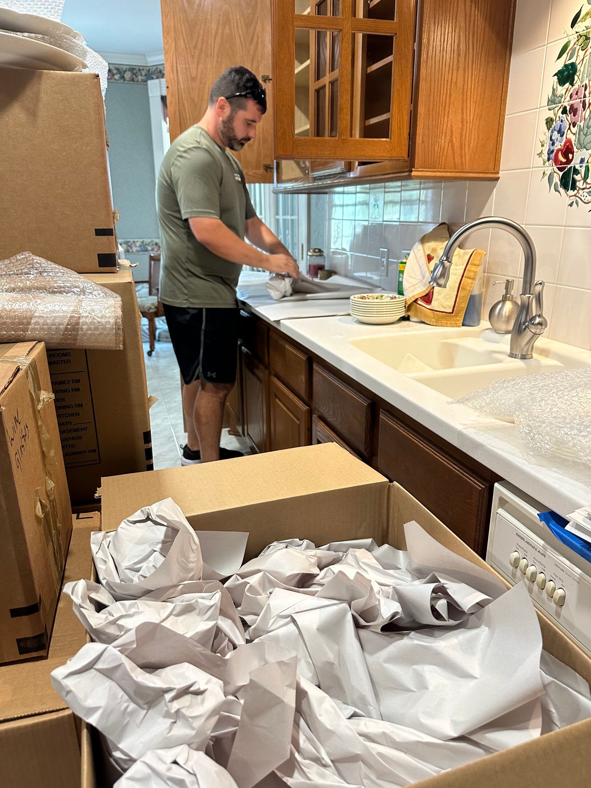 A man is standing in a kitchen surrounded by boxes.