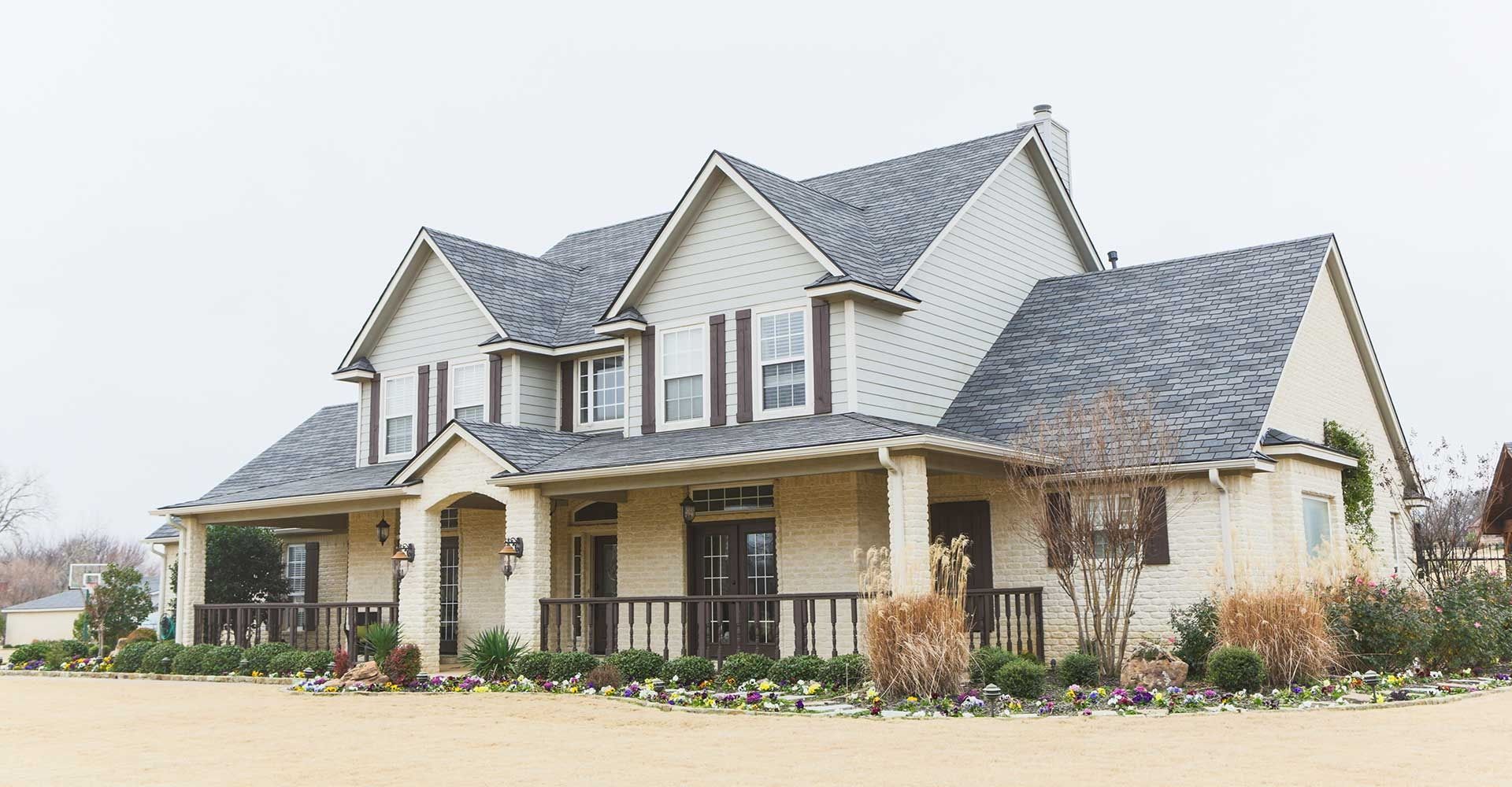 Large two-story suburban house with gray gabled roof and beige brick exterior on a landscaped lot