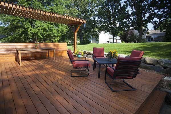 Wooden deck patio with red chairs and a glass table under a pergola, surrounded by trees and landscaping