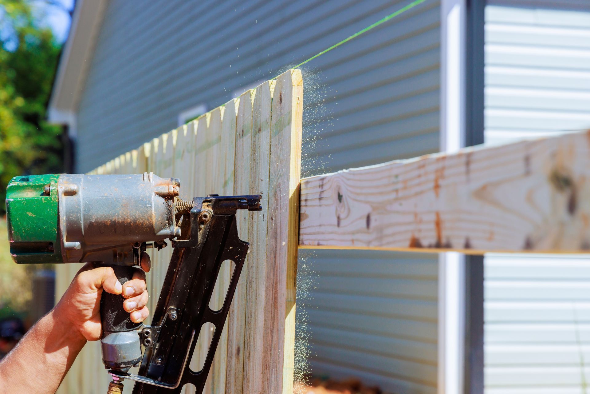 Hand holding a nail gun fastening a wooden board to a fence in a sunny yard