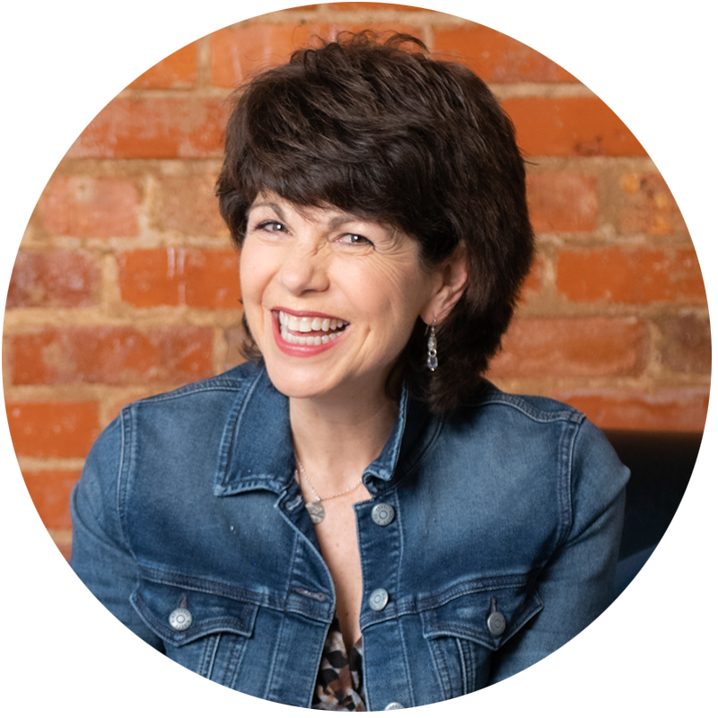 A woman in a denim jacket is smiling in front of a brick wall