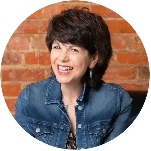 A woman in a denim jacket is smiling in front of a brick wall