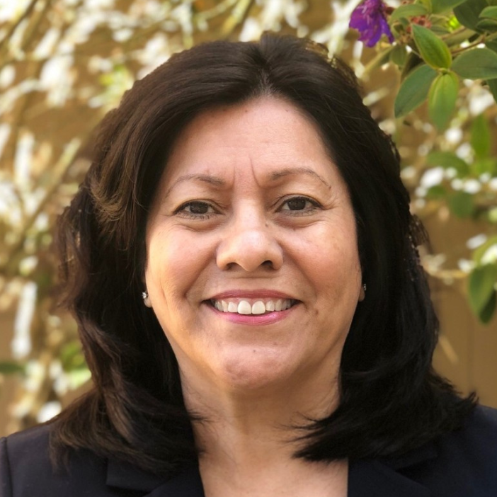 A woman with dark hair is smiling in front of purple flowers