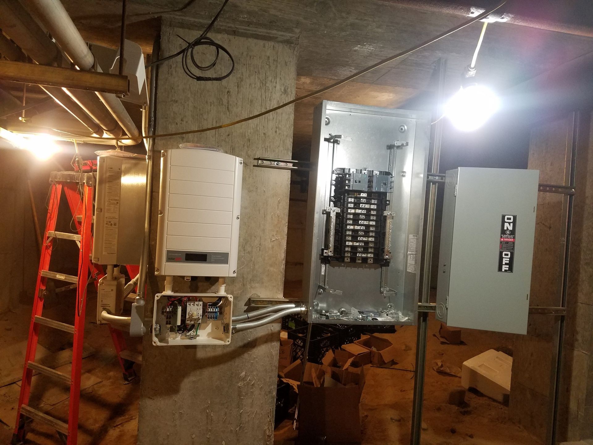 Electrical panel installation in a basement; breaker boxes, conduit, ladder, and work lights.