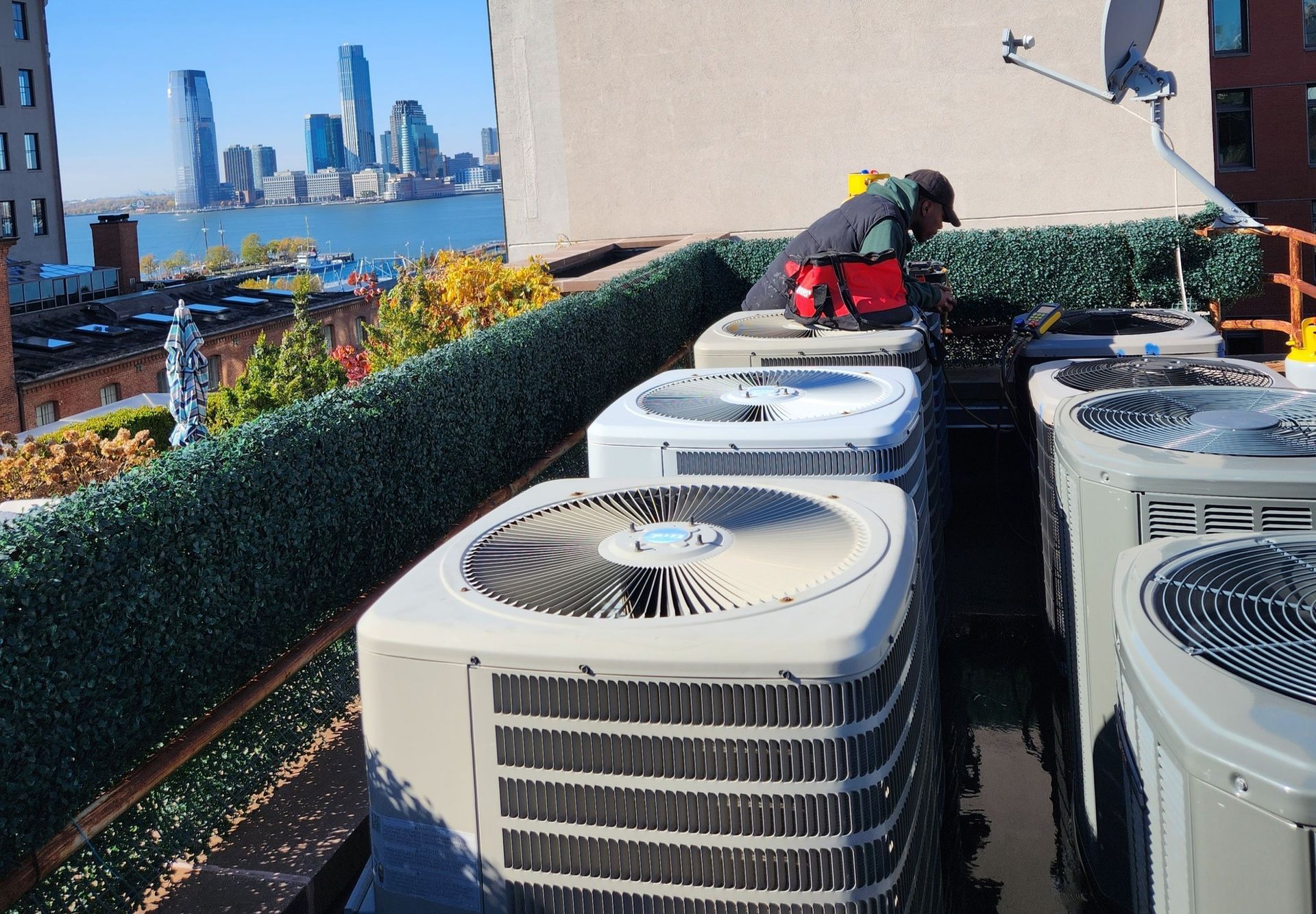 Rooftop with HVAC units; a person in work clothes inspects the equipment with a cityscape in the background.