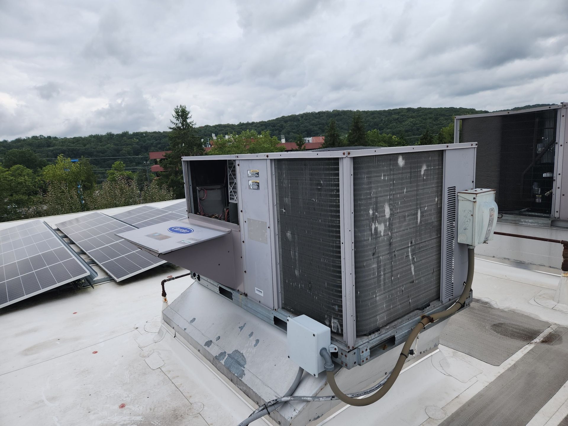 HVAC unit on a rooftop with solar panels, facing a green hillside under a cloudy sky.
