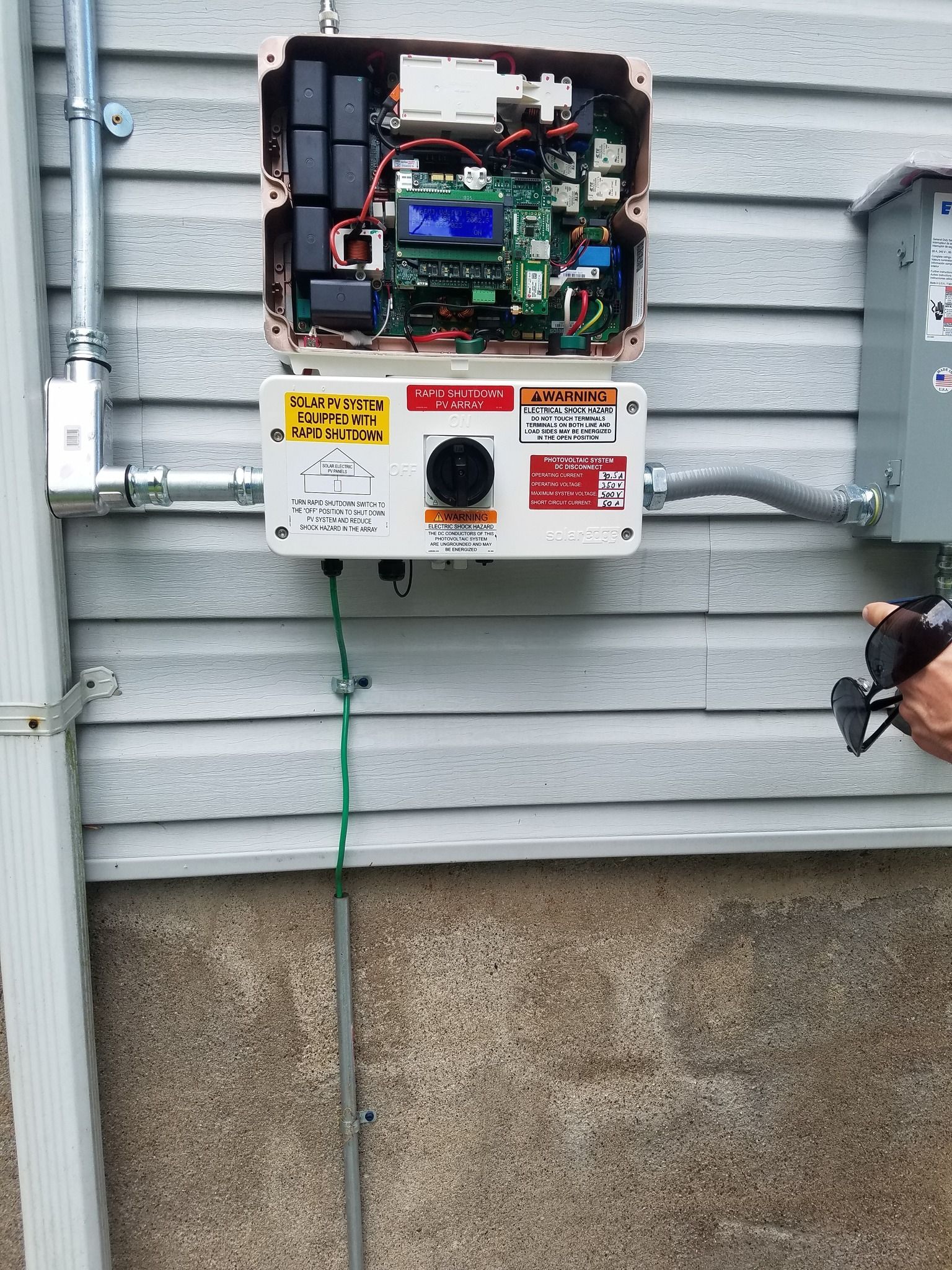 Solar panel inverter box mounted on a gray siding wall, connected to conduits and a gray electrical box.