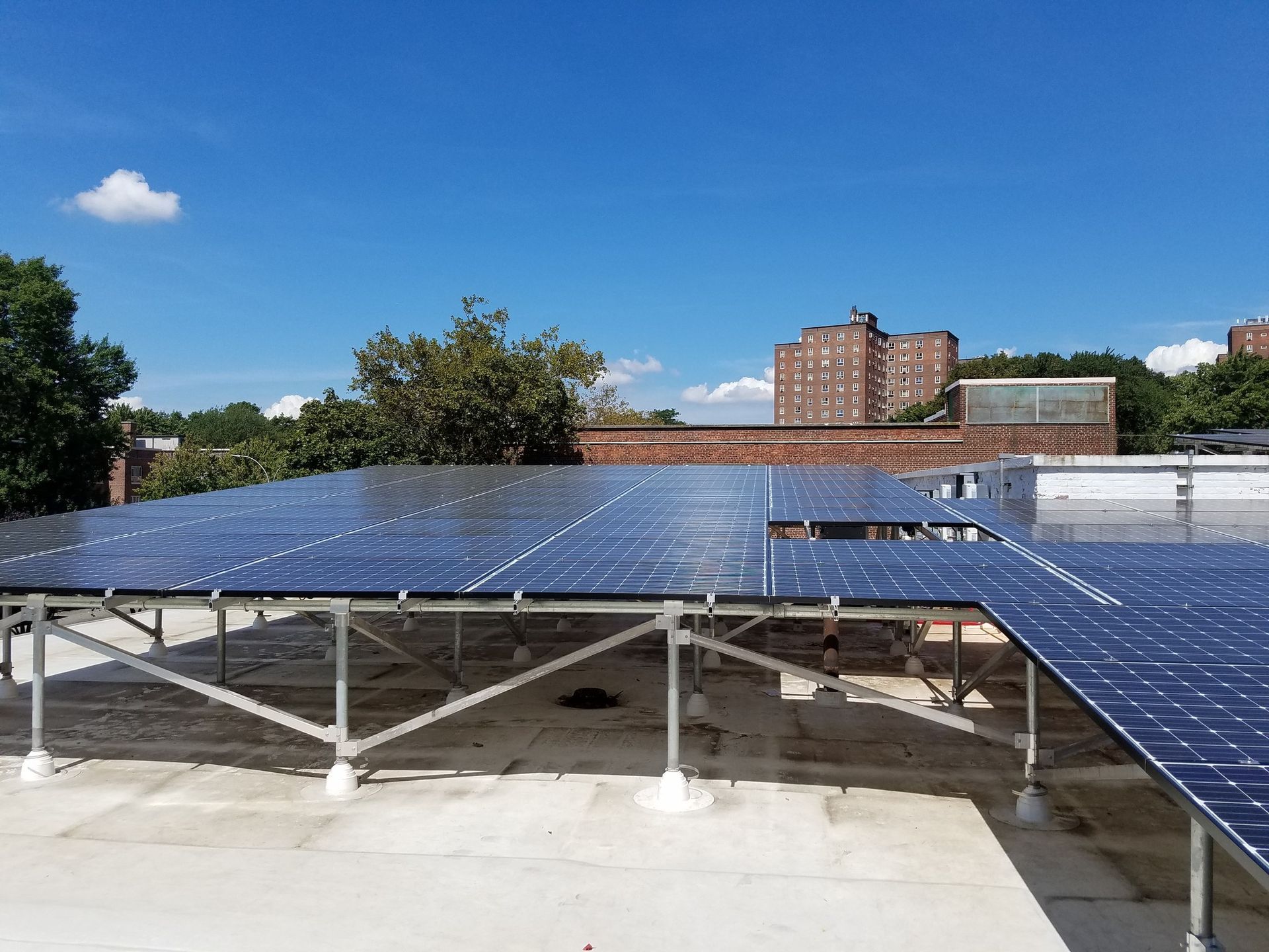 Solar panels installed on a rooftop against a blue sky, with buildings and trees visible.