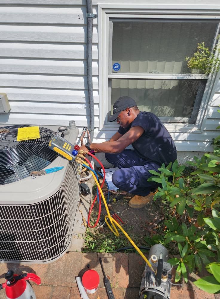 HVAC technician working on an AC unit outdoors; connecting hoses with gauge, next to a window.