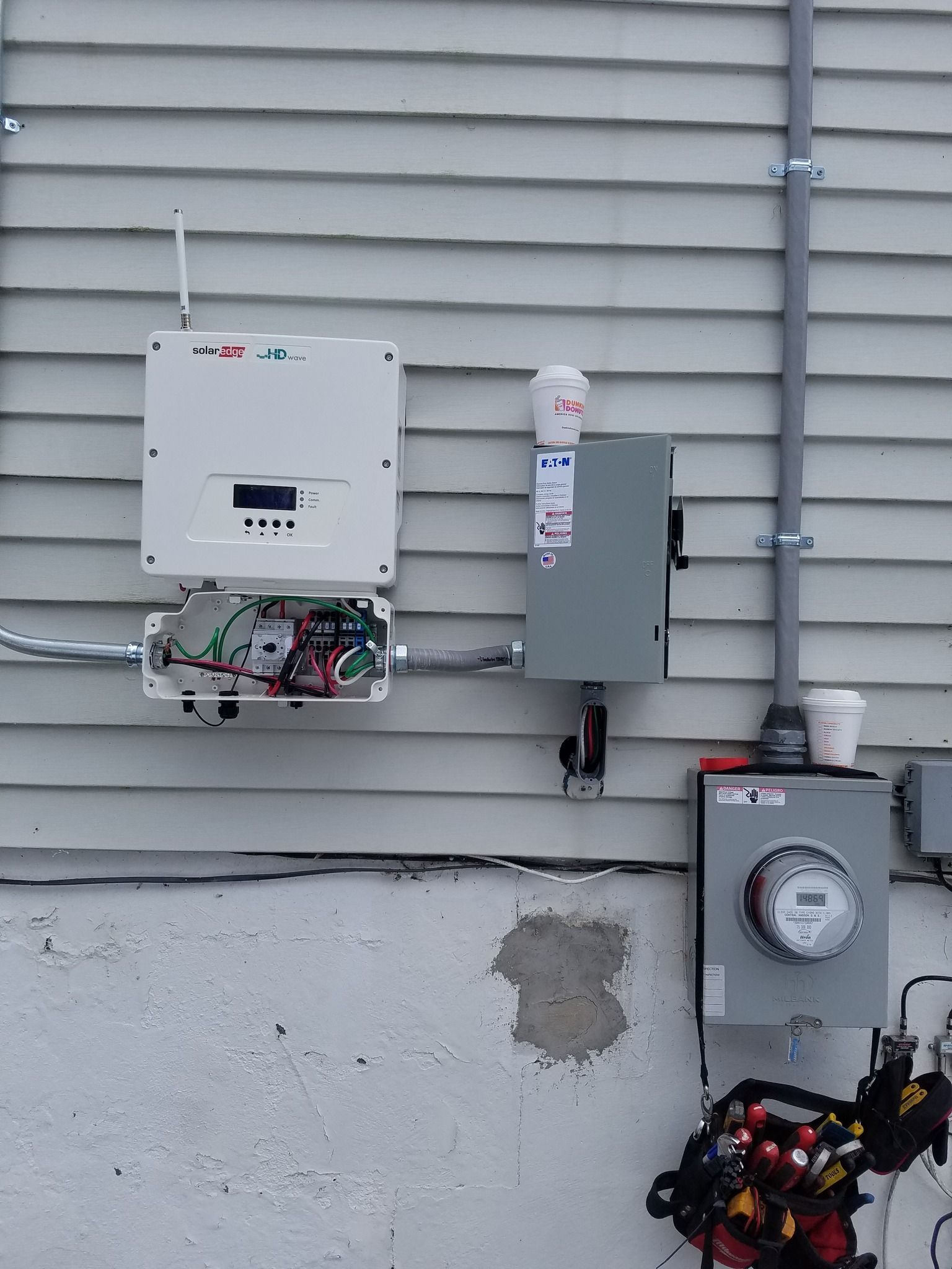 Solar panel equipment mounted on a white house exterior with a meter, conduit, and inverter.