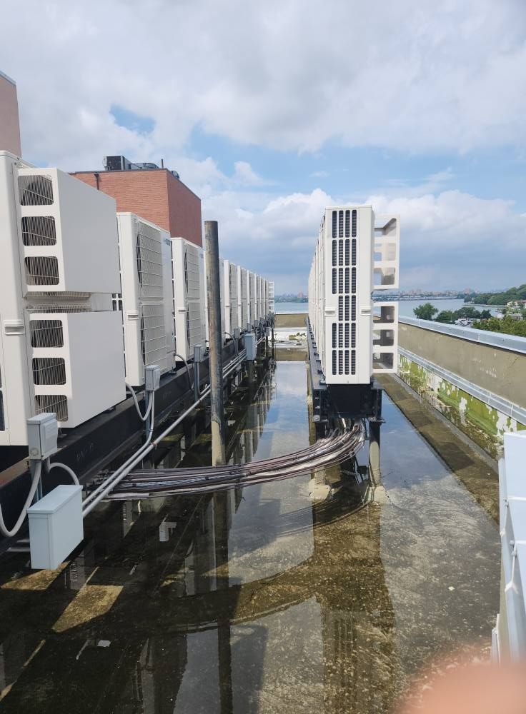 Rows of white air conditioning units on a rooftop with standing water and an overcast sky.